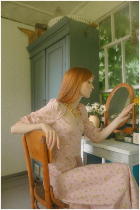 Woman in pink floral dress sits at vanity, wearing