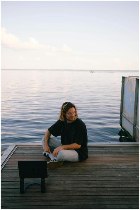 A man with long hair sits on a pier by the sea usi