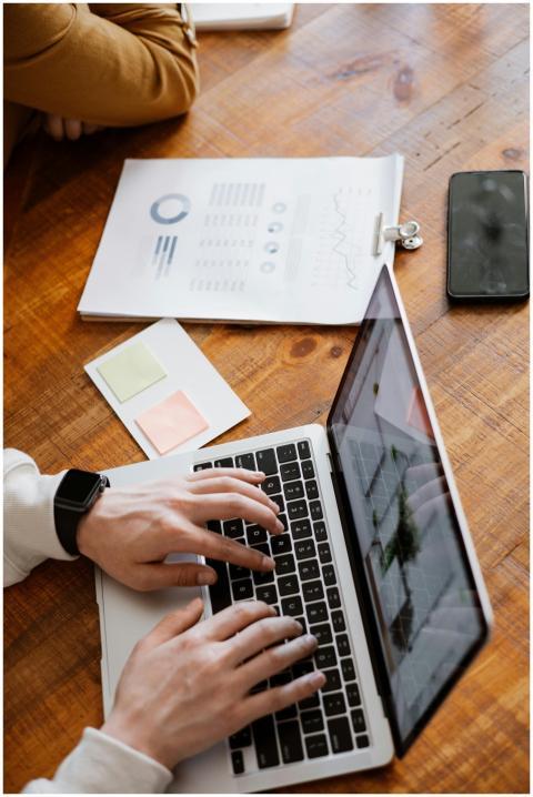 Hands typing on a laptop with paperwork and smartp