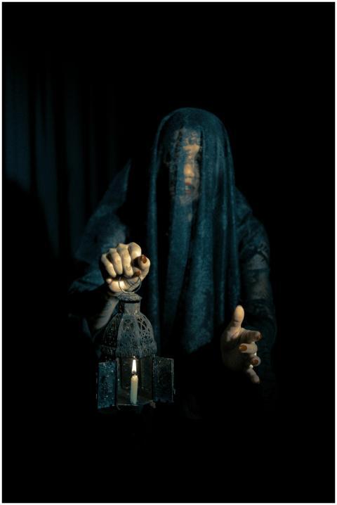 A cloaked figure holds a lit lantern in an enigmat