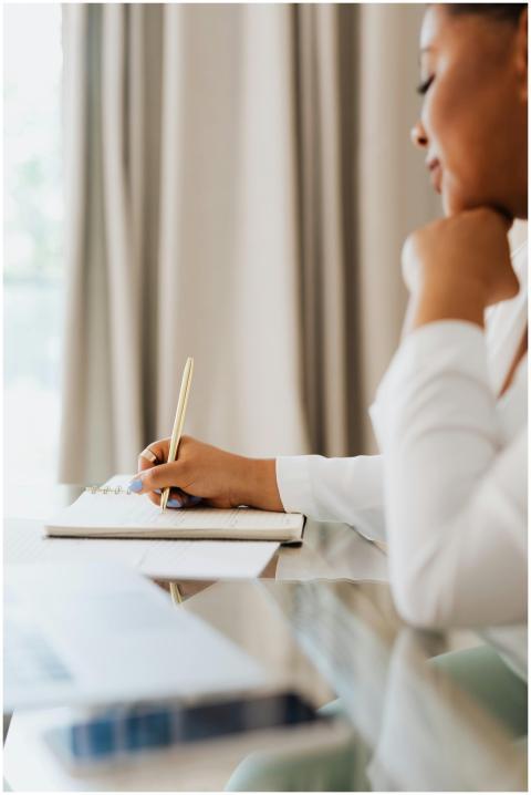 A woman sits indoors writing notes in a notebook w
