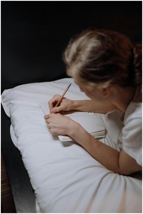 Woman lying on bed writing in a notebook, dimly li