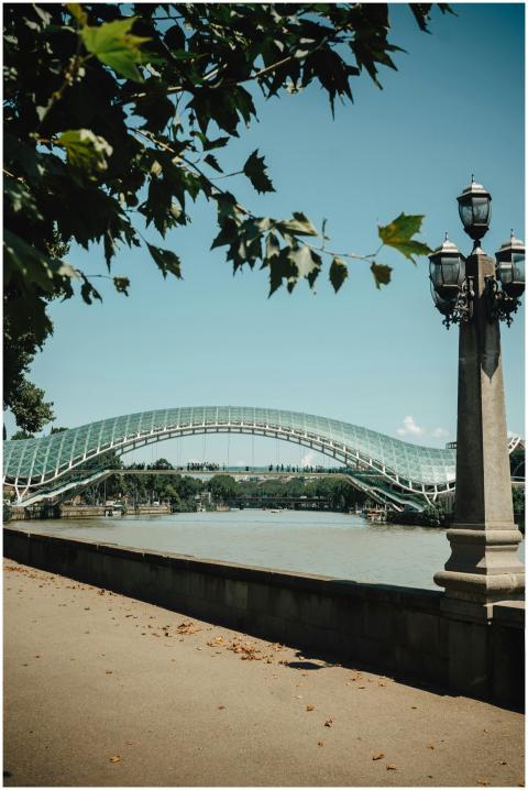 A modern glass bridge spans a river in an urban pa