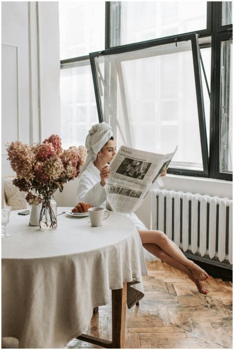 Woman in bathrobe reading newspaper by window, enj