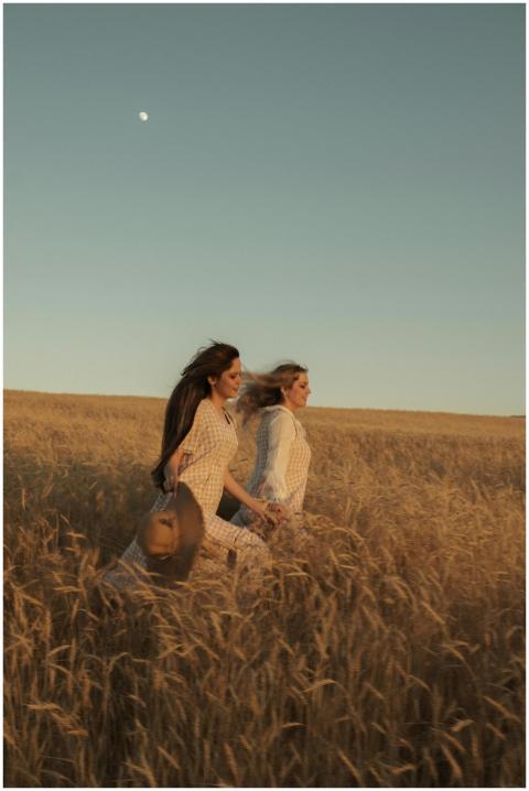 Two women run through a sunlit wheat field under a