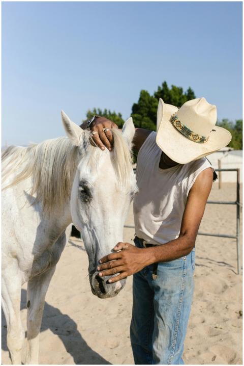 A cowboy in a hat gently strokes a white horse at