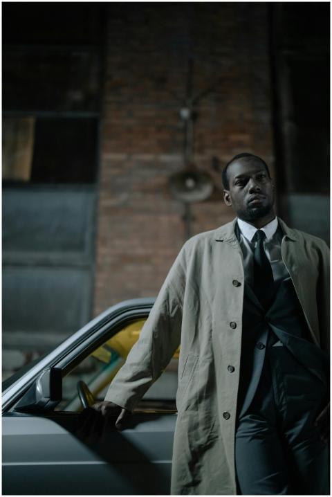 A stylish man in a trench coat leans on a car in a