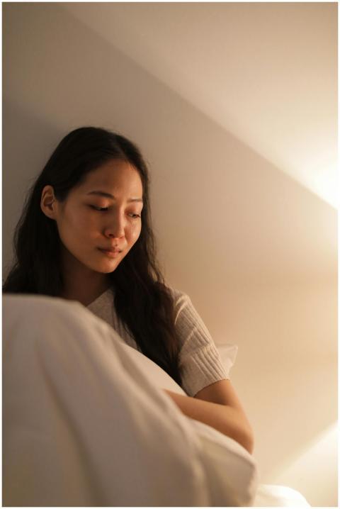 A young woman sits thoughtfully in a cozy bedroom,