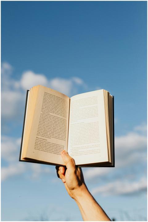 A hand holding an open book against a blue sky; sy