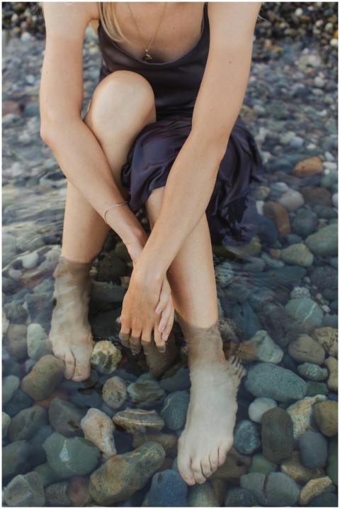 A woman sits with her feet in rocky beach waters,