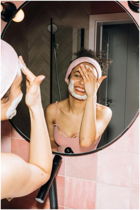 Happy woman applying face mask while smiling in ba