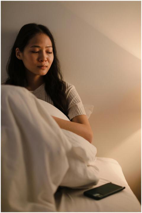A thoughtful Asian woman sits on a bed, wrapped in