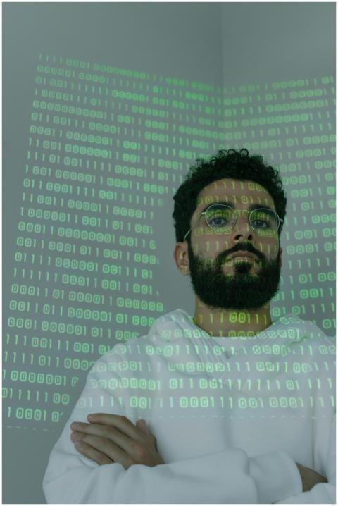A bearded man in glasses stands with binary code p