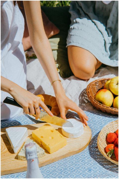 A sunny picnic scene with hands cutting cheese, su