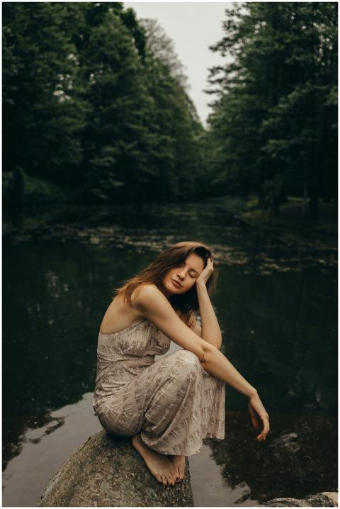 A woman in a dress sits peacefully by a calm river