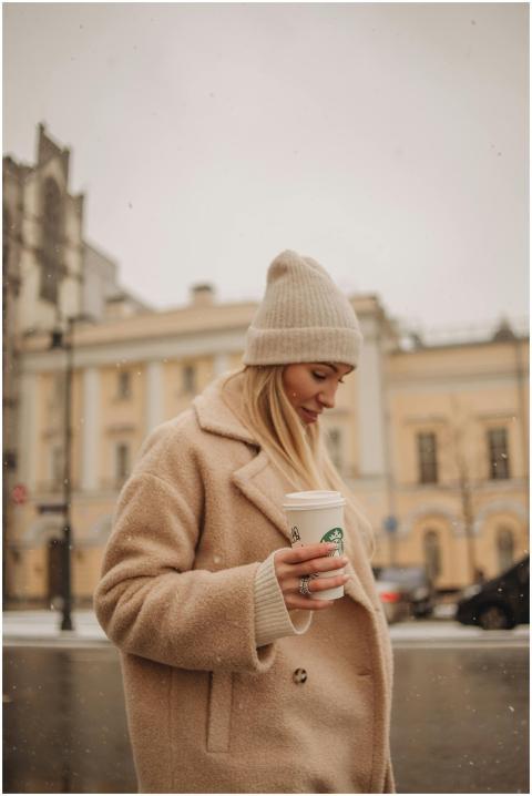 A woman in a beige coat holds a cup of coffee whil
