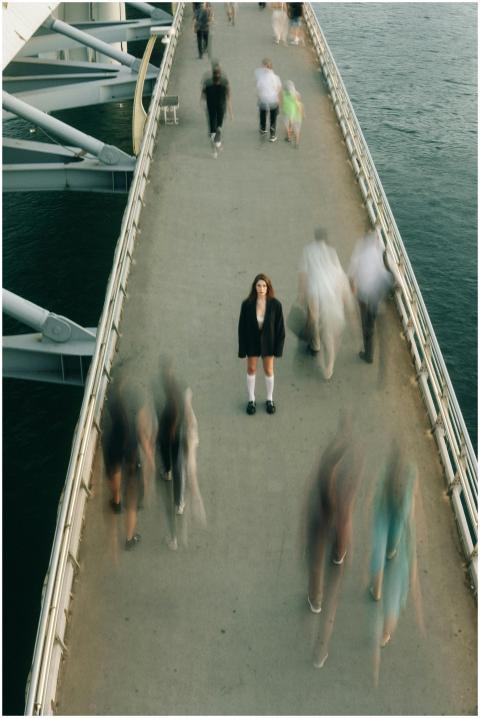 A woman stands still on a bustling footbridge, cap