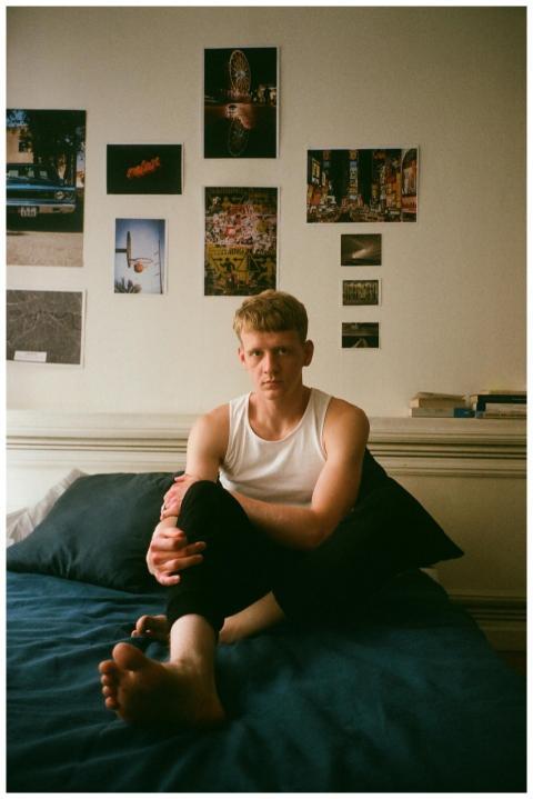 A young man in a tank top sits on a bed in a creat