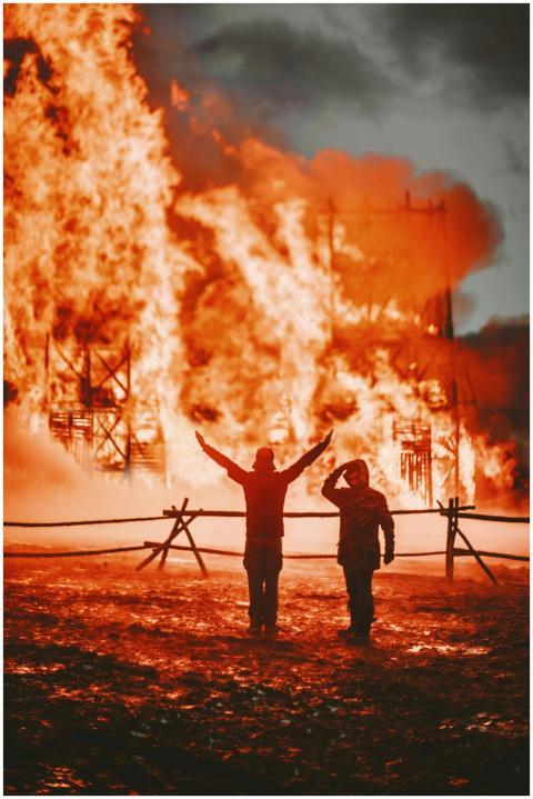 Silhouetted figures stand against a massive blaze,