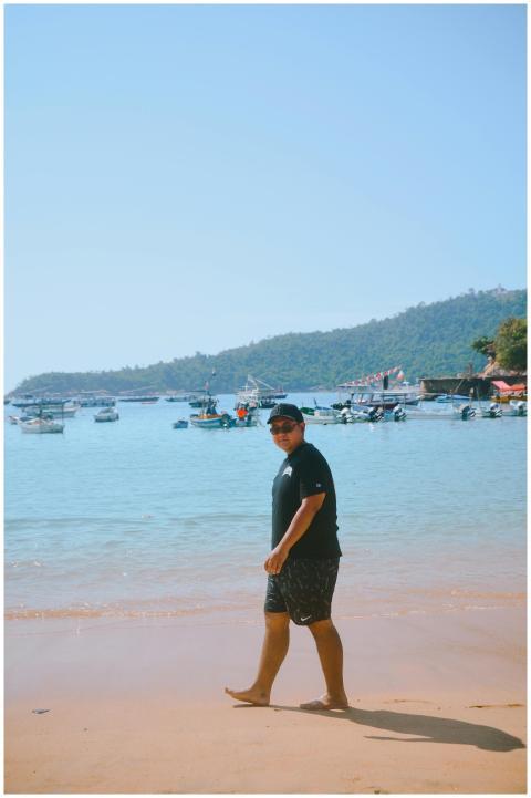 A man strolls along a sunny beach with boats ancho
