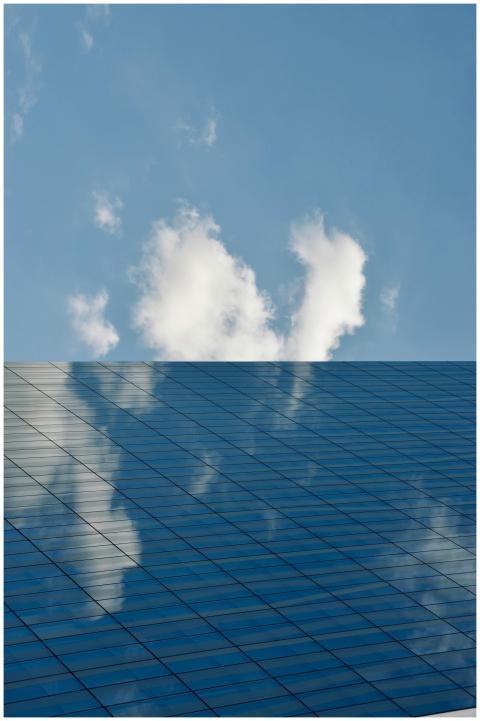 A glass skyscraper reflects clouds and blue sky, c
