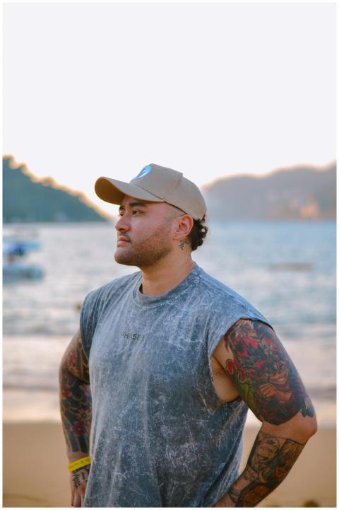 A tattooed man stands on a sandy beach, enjoying a