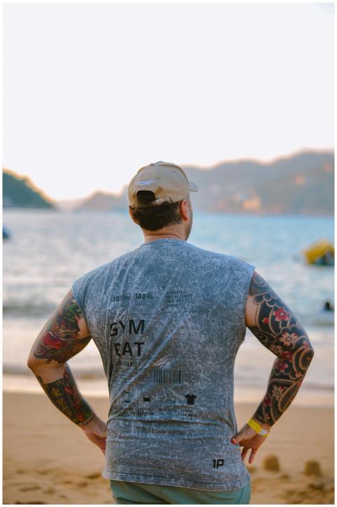 A tattooed man stands on a sandy beach, gazing at