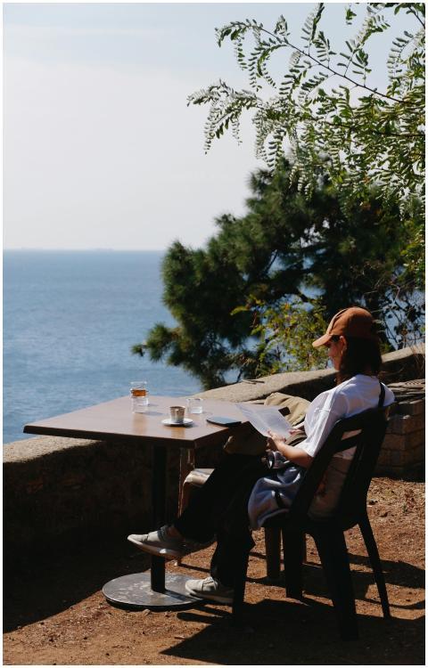 Person reading at an outdoor table by the sea, enj
