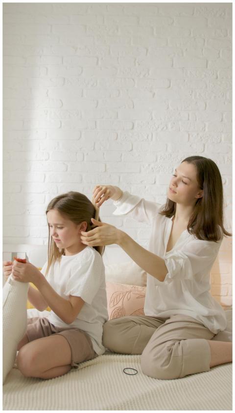 A mother gently combs her daughter's hair on a bed
