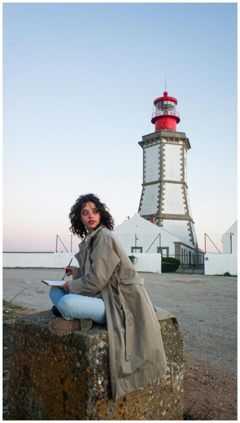 A woman in a trench coat sits near a lighthouse du