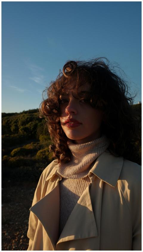 A woman with curly hair poses outdoors in a beige