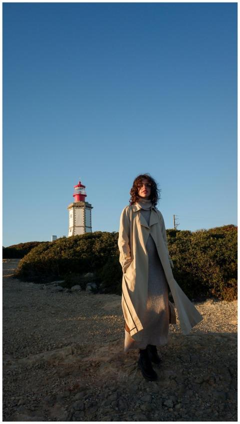 A woman in a beige coat stands near a lighthouse w