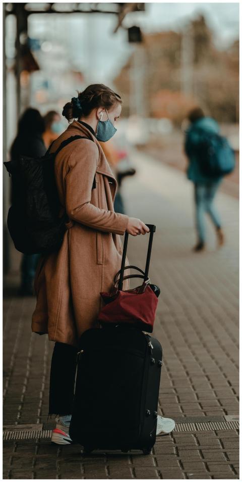 A woman in a coat and mask waits with luggage at a